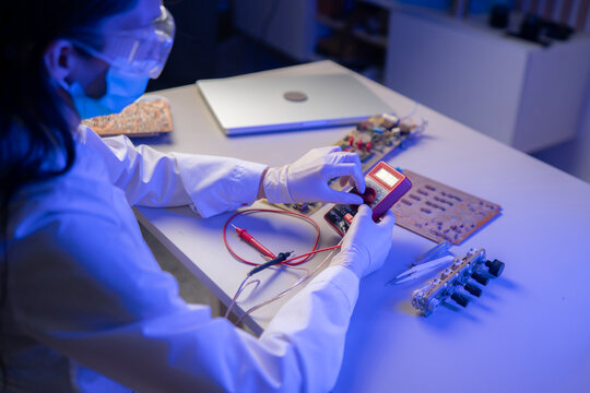 A person in a lab coat tests a circuit with a multimeter while wearing gloves and a mask in a workspace