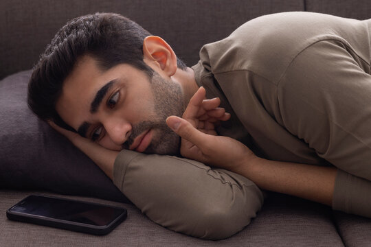 Portrait of a young man lying on sofa and looking at the Smartphone kept beside 