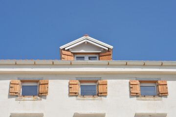 Obraz premium A low angle view captures a white plaster house facade featuring three small windows with open light brown wooden shutters and a dormer above against a clear blue sky