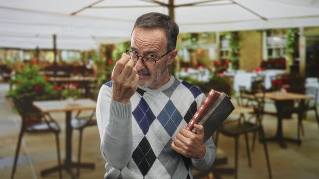 Man holding books and making pinched fingers gesture in street cafe patio under umbrellas; quiet curiosity.