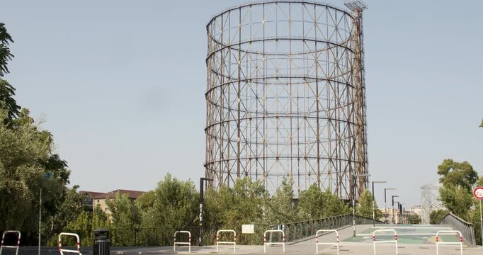 A wide shot of a industrial gasometer metal framework rising above green trees, modern street barriers, and a paved walkway under a clear, bright sky on a warm sunny day. It's located in Rome, Italy.