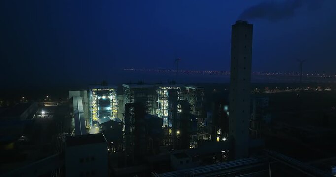 Industrial power plant operating at night with glowing lights and a tall chimney stack under a dark blue sky.