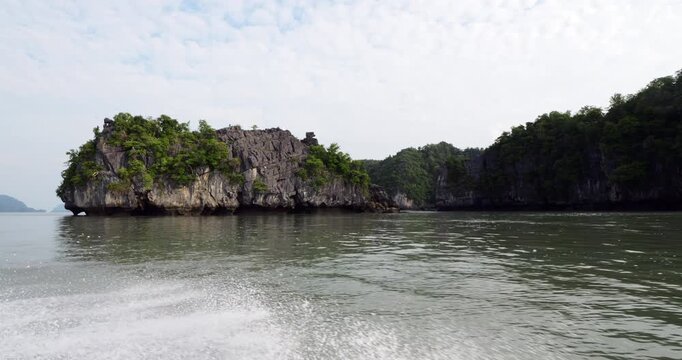 Slow motion side view from tourist boat passing rugged limestone islet at seaward edge of Kilim Karst Geoforest Park. Tropical greenery clings to rough black rocks, calm waters of Strait of Malacca
