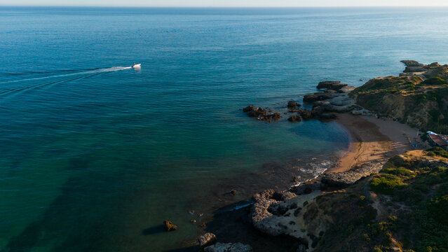 aerial view of boat sailing on rocky coastline with sandy beach under clear blue sky, relaxing and serene scene for travel and vacation backgrounds, clean and modern design Praia dos Aveiros Portugal