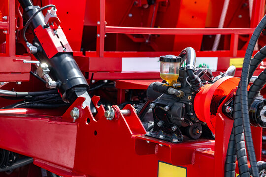 Agricultural machinery hydraulic fluid pump with oil reservoir. Modern farming equipment red metal frame chassis assembly with piston cylinder and industrial transport engineering details.