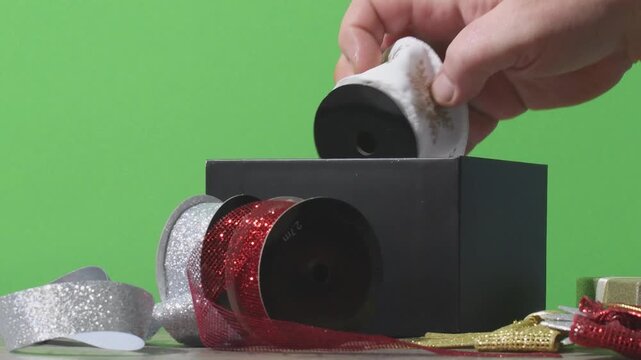 Hands place colorful gift wrapping ribbons back into a box after preparing for a present. The setting shows a clear background with wrapping materials and presents nearby.