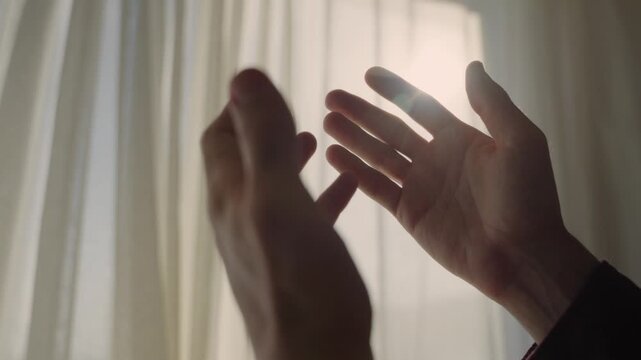 Man's hands clasping together in a praying gesture against a bright window with curtains