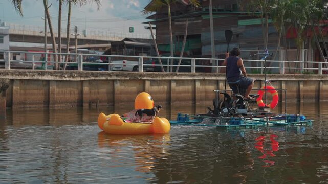 Man riding pedal catamaran towing dog on inflatable duck through calm canal, Pet doggy enjoying river ride behind floating boat 4k