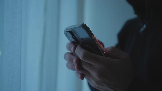 Close up on hooded person in dark room using mobile phone texting and sending information. Illegal or shady business activity in closed hidden room. Unrecognized man holding smartphone. Spy or hacker