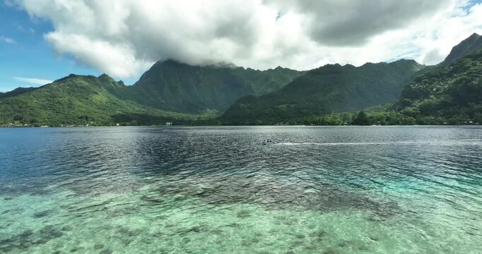 Jagged green mountain peaks rise above calm lagoon waters in French Polynesia, faint boat wake trails across rippling water beneath dramatic thick storm-cloud covered sky. Aerial drone view.