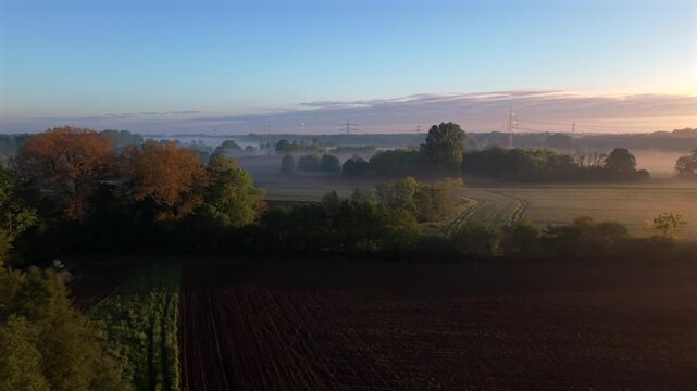 Freshly tilled dark soil field stretches beside golden autumn tree line in rural USA, thin morning fog drifts across misty farmland beneath soft pastel sunrise skies. Aerial drone wide shot