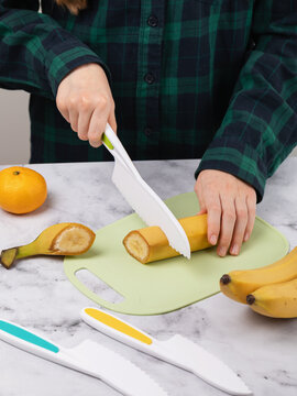 Child girl cutting banana with kids safe plastic knife on cutting board in kitchen faceless