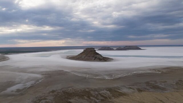 A stunning cinematic drone shot of a single dramatic rock outlier standing isolated in the shallow waters of Karynzharyk depression, surrounded by a soft, milky-white haze in Mangystau, Kazakhstan