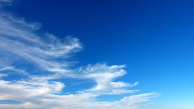 Wide shot of thin wispy white cirrus clouds scattered across a vast clear blue sky on a bright sunny day, atmospheric cloud patterns drifting slowly in the high altitude atmosphere.