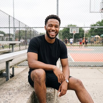 Happy young Black man in 20s smiling while sitting on bleachers at outdoor basketball court during daytime, looking at camera, fitness concept