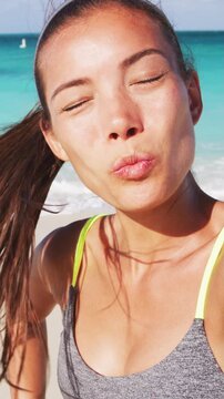 Woman on beach blowing kiss having fun on beach vacation during summer travel holidays taking a selfie photo or having video chat. Vertical video of real unretouched woman model without any makeup