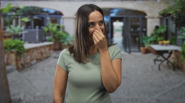 Woman covering mouth with hand, smiling and laughing near a stone building courtyard entrance with potted plants and table; amusement warmth.