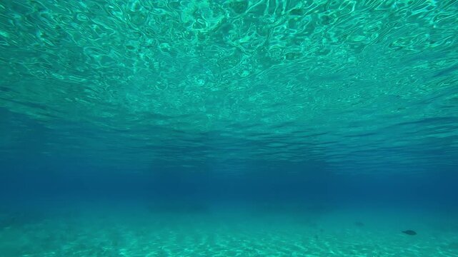 Emerald green water surface with fine ripples above sandy seabed on sunny day, underwater view. Turquoise water with emerald surface over sandy shallow water and sun glare on the sand