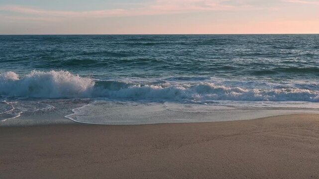 Gentle ocean waves roll onto the sandy beach as the warm golden light of the setting sun reflects beautifully across the water&rsquo;s surface