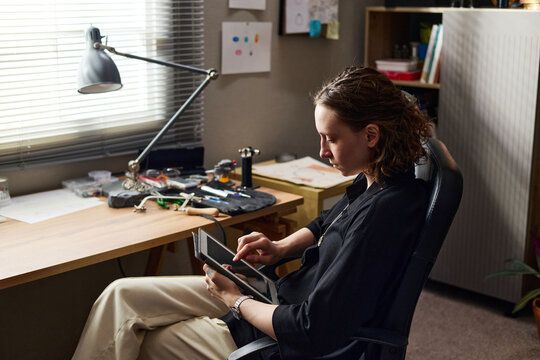 Young adult Caucasian female jeweler using tablet while sitting at home desk, managing remote workflow. Ideal for remote work, productivity, freelance business themes