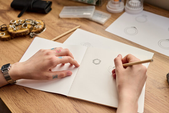 Close up of young adult woman sketching jewelry designs in notebook on desk, developing handmade product collection. Ideal for artisan branding, ecommerce, creative business