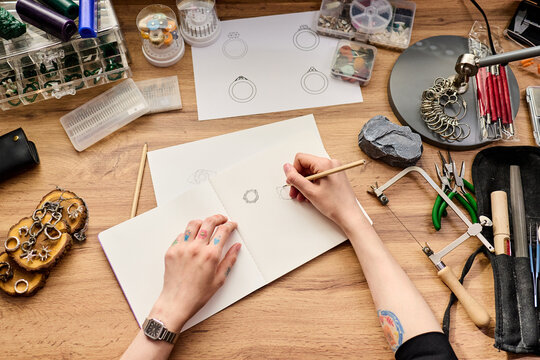 Top view shot of young adult woman sketching ring designs in notebook at jewelry workbench, planning handmade product collection. Ideal for artisan branding, ecommerce, craft business