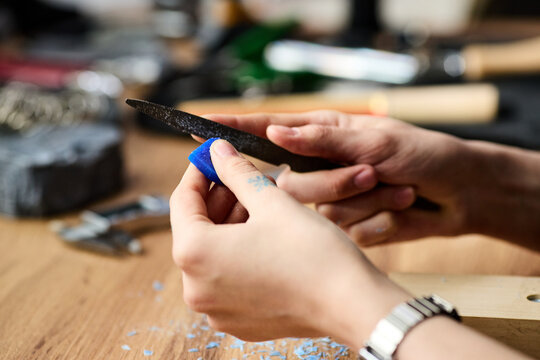 Close up of young adult hands filing blue wax piece at workbench, shaping craft material for handmade production. Useful for artisan business, maker workshop, product process marketing