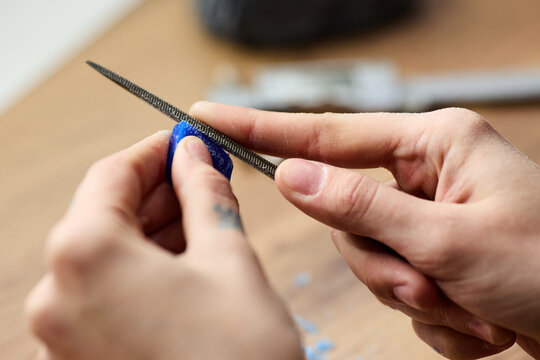 Close up of young adult hands filing small blue object on workbench, shaping handmade product with precision. Suitable for craft business, artisan branding, DIY tutorial content