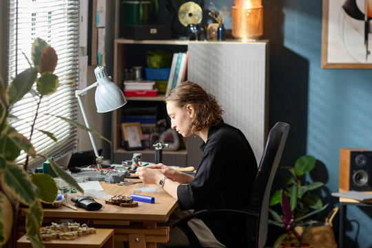 Young adult Caucasian woman crafting jewelry at workbench, assembling handmade accessories in home studio. Ideal for artisan business, creative entrepreneurship, small brand marketing