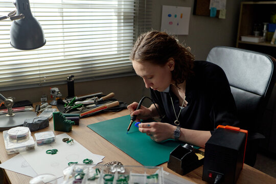Young adult Caucasian woman crafting jewelry at workbench, using precision tool on handmade pieces for artisan production. Ideal for small business, creative entrepreneurship marketing