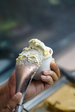 Fresh artisan gelato served into a white paper cup using a metal scoop. Authentic preparation process with shallow depth of field, ideal for hospitality, dessert and food service concepts.