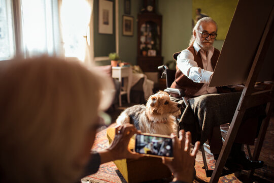 Senior man painting at home while woman photographs and dog watches