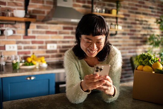 Smiling woman using smartphone in home kitchen with fresh groceries