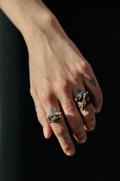 Vertical close up shot of young adult womans hand displaying multiple rings and small finger tattoos against black background. Supporting jewelry branding, accessory retail, style campaign visuals