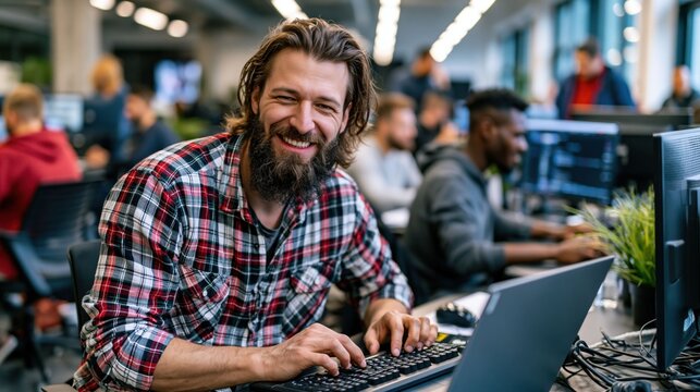 man with a beard is sitting at a desk using a laptop. He is smiling at camera while others work on their computers in a busy office. Light comes in from large windows