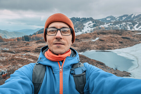 Man smiling at camera, capturing a selfie during a mountain hike with a partially frozen glacier lake and autumn hills
