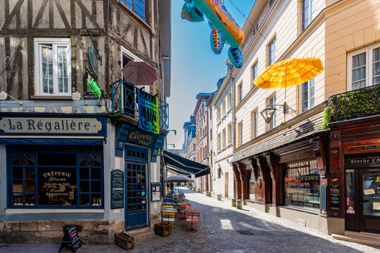 Wide sunny view of the corner of Rue Massacre in Rouen, France, showing the La R&eacute;gali&egrave;re and La Cr&ecirc;perie Bleue storefronts with colorful bistro chairs, inflatable sea turtle and yellow raffia parasol 