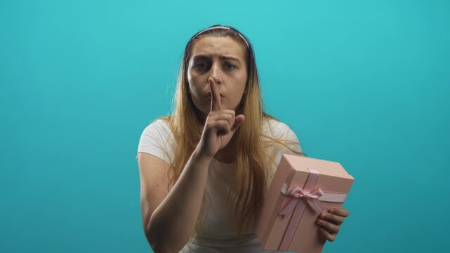 Woman holding a pink present and pressing finger to lips in studio, leaning forward with worried glance and tense expression; secret silence.