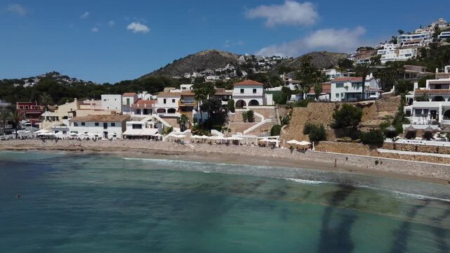 Cinematic drone shot approaching the turquoise waters and scenic shoreline of El Portet cove in Moraira, Spain, showcasing a sunny Mediterranean beach lifestyle from an aerial view.