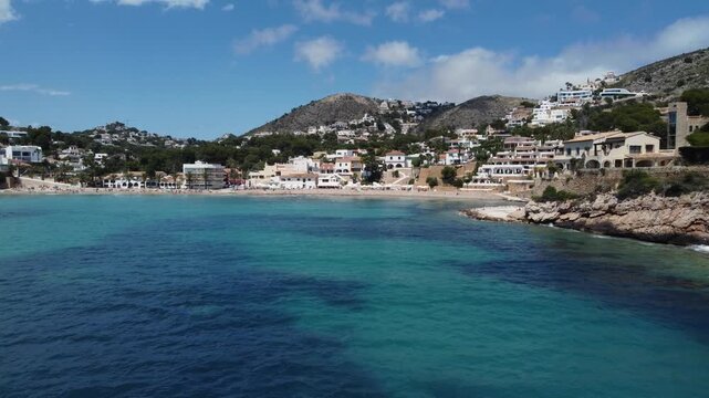Drone footage flying fast and low over the sea toward El Portet beach in Moraira, Spain, passing an anchored boat on a sunny summer day.