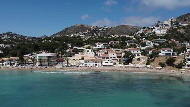 Ascending drone flight reveals a stunning aerial view of El Portet beach in Moraira, Spain, showcasing crystal-clear turquoise waters and rugged coastline.