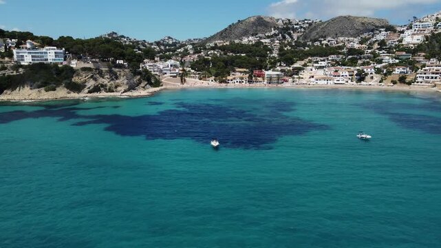 Cinematic drone shot flying sideways along luxury boats anchored in the turquoise waters of El Portet beach, Moraira, Spain, showcasing a sunny Mediterranean vacation vibe.
