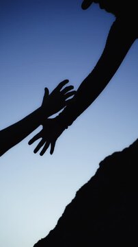 Dramatic silhouette of helping hand pulling another climber up steep mountain rock, symbolizing assistance, support, trust, teamwork, and achieving goals together