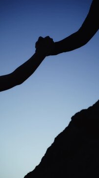 Dramatic silhouette of person extending hand to help female hiker ascend steep mountain rock against  clear blue sky, illustrating assistance, partnership, and achievement