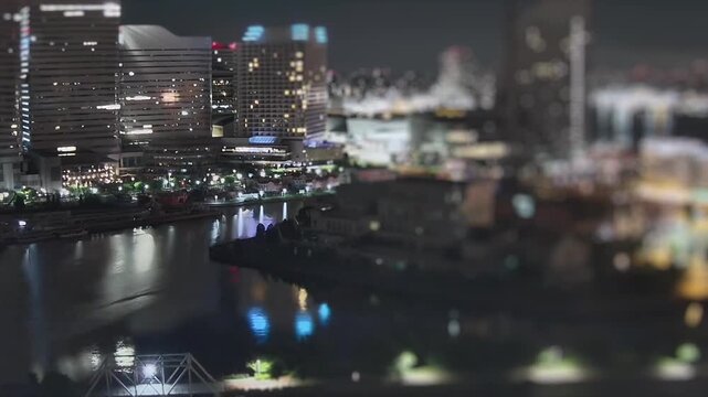 Night timelapse of Yokohama waterfront in Japan with illuminated high rises and reflections on the harbor canal. Tilt shift style cityscape with glowing skyline.