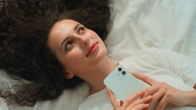 Top view pensive Caucasian woman smiling thinking relaxing holding mobile phone lying in bed positive dreamy female girl with smartphone application telephone gadget resting think at morning bedroom