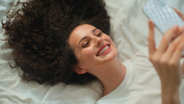 Smiling carefree Caucasian woman using mobile phone network technology enjoying online shopping lying on bed female girl with smartphone smile texting with messenger relaxing on linen white bedsheets