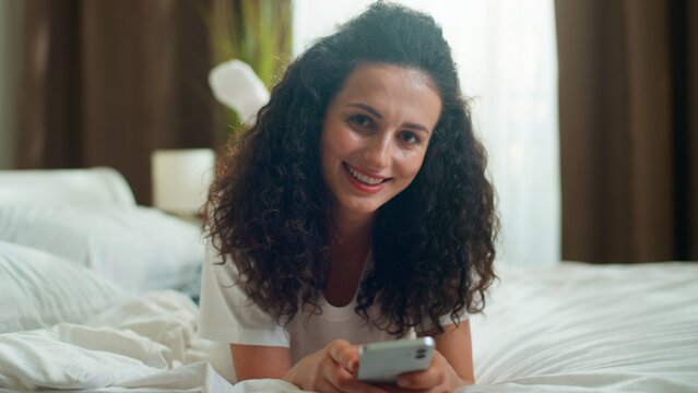 Caucasian relaxed calm woman using mobile phone app smiling in bedroom at home happy girl female browsing smartphone social media chatting smile looking at camera relaxing at domestic weekend at bed