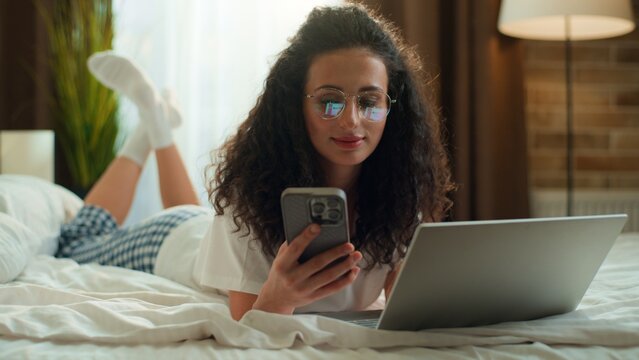 Caucasian woman using laptop and mobile phone lying relaxing in bedroom on comfortable bed smiling happy female girl browsing computer multitasking with smartphone optimistic gadget addict at morning