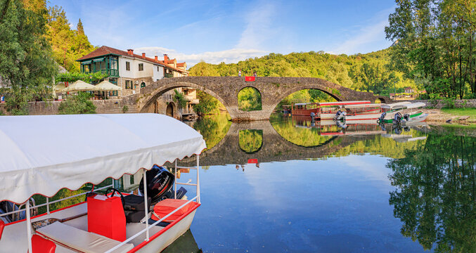 25 May 2026, Rijeka Crnojevica, Montenegro: Old stone bridge arching over calm river, Tourist boats are awaiting passengers in this historic Montenegrin village